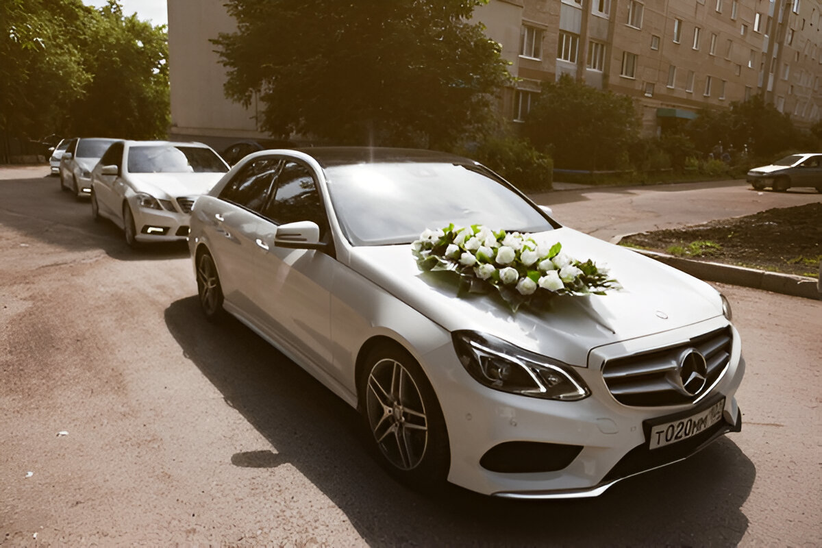 Düğün için süslenmiş beyaz Mercedes, ön kaputunda beyaz çiçek aranjmanı ile önde ilerliyor.